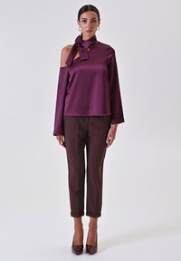 Top in satin bordeaux con un fiocco al colletto, con design off-shoulder; abbinato a pantaloni scuri a sigaretta e tacchi alti.