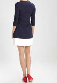 Marineblaues Kleid mit drei Viertelärmeln und weißem Saum, glattes Material, taillierte Passform und Reißverschluss in der Mitte des Rückens. Getragen mit roten Schuhen.