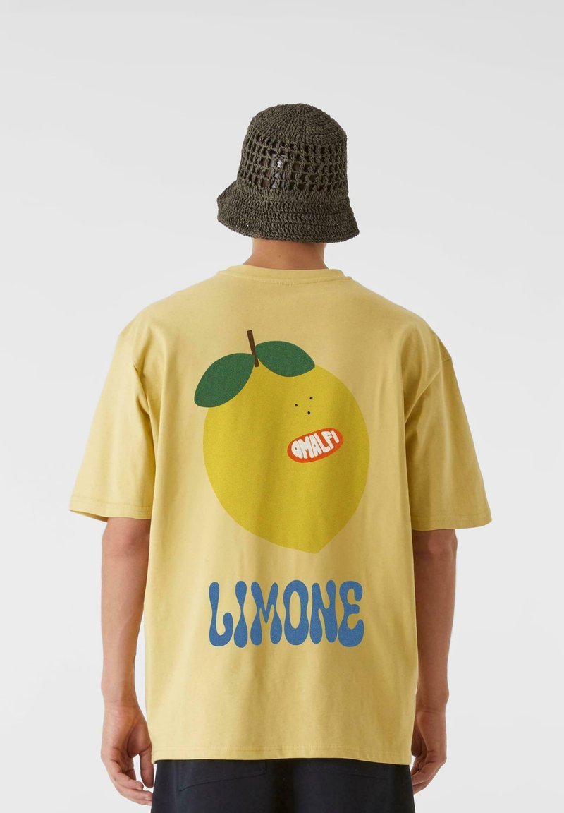 Gul bomull T-skjorte med et stort sitronmotiv og teksten "LIMONE" i blått. Komplimenterer en teksturert olivengrønn bøttehatt.