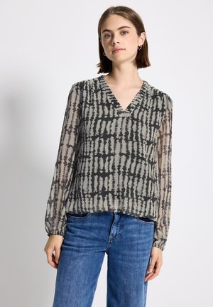 Une jeune femme portant un chemisier transparent à manches longues, noir et gris à motifs, et un jean bleu, se tenant devant un fond blanc uni.
