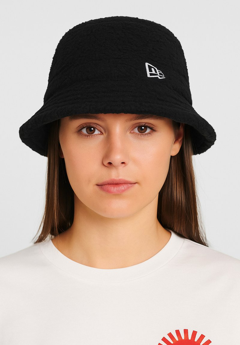 Schwarzer Bucket Hat mit strukturierten Oberflächen und einem kleinen weißen Logo auf der Vorderseite, getragen mit einem schlichten weißen T-Shirt, das ein rotes Grafikdesign zeigt.