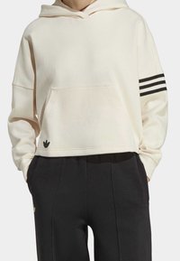 Kremowa, krótka bluza z kapturem wykonana z miękkiego materiału, z przednią kieszenią i trzema czarnymi paskami na lewym rękawie. Czarny detal z logo na dole.
