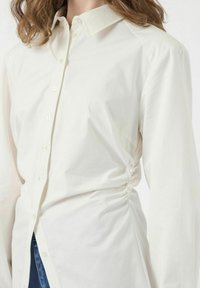 Witte katoenen button-up shirt met lange mouwen, voorzien van geplooide details aan de zijkanten en een licht oversized pasvorm. Soepele textuur en klassieke kraag.