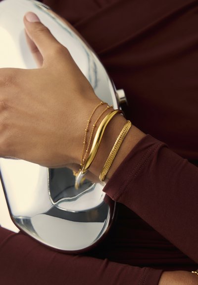 Złote bransoletki o gładkich, skręconych i plecionych wzorach noszone na nadgarstku na maroonowym tle z odbiciami.