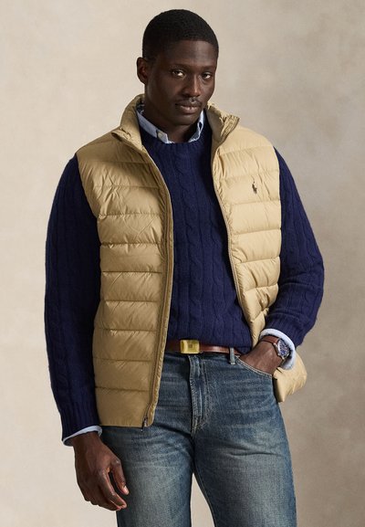 Doudoune sans manches beige, pull en tricot bleu marine et jean bleu. La doudoune présente une texture matelassée et un col montant. Accessoires : une montre et une ceinture.