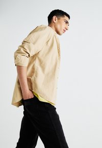 Beige overhemd met knopen en opgerolde mouwen, gedragen over een gele shirt, gecombineerd met zwarte jeans. Glad materiaal en casual ontwerp.