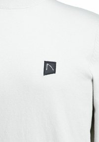 Witte gebreide trui met een ribbelstructuur ronde halslijn en een klein zwart patch met witte details. Zachte textuur met een slim-fit vorm.