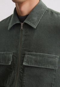 Zielona kurtka sztruksowa z zamkiem z przodu, ostrym kołnierzem oraz dwoma kieszeniami na piersi z klapami. Teksturowana powierzchnia z pionowymi paskami.