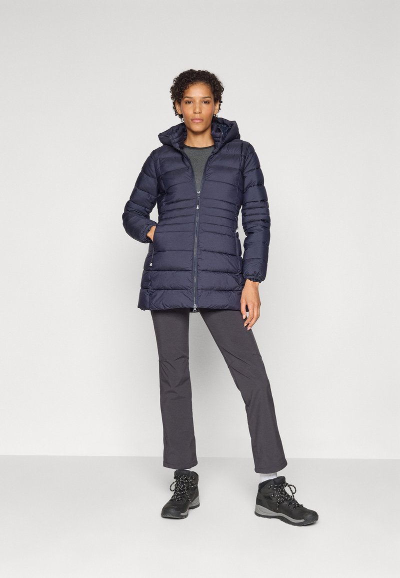 Giubbotto imbottito blu navy con design aderente, chiusura con zip e cappuccio; presenta cuciture verticali e tasche laterali. Pantaloni neri e scarponi da trekking completano l'outfit.