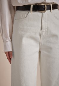 Weiße Jeanshose mit lockerer Passform, ausgestattet mit einer Vordertasche und einem schwarzen Ledergürtel mit einer silbernen dekorativen Schnalle.