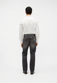 Άνδρας που φαίνεται από πίσω, φοράει λευκό πουκάμισο με μακριά μανίκια, ξεβαμμένα μαύρα τζιν με πίσω τσέπες και σκούρα γυαλισμένα παπούτσια, σε λευκό φόντο.