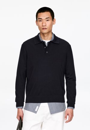 Jonge man die een zwarte polotrui met lange mouwen draagt over een gestreept overhemd, witte broek, en een zwarte tas vasthoudt tegen een witte achtergrond.