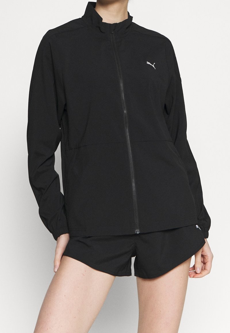 Chaqueta ligera negra con cremallera frontal, cuello alto y puños elásticos. Presenta el logo de Puma en el lado izquierdo del pecho. Combinada con pantalones cortos negros.