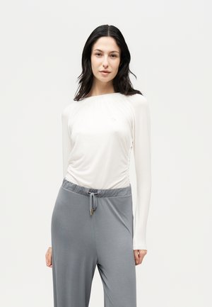 Une femme porte un haut blanc à manches longues et un pantalon gris à cordon de serrage sur un fond uni et clair.
