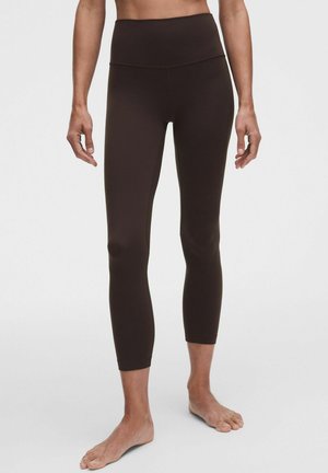 Kvinde iført mørkebrune cropped leggings med højtaljet pasform, stående barfodet mod en ensfarvet lys baggrund.