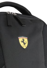 Sac à dos noir avec un tissu texturé, des accents en maille, des double zip, et un logo Ferrari sur un écusson en forme de bouclier.