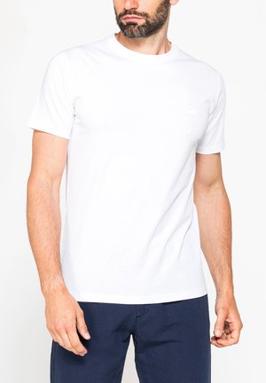 Uomo con barba che indossa una maglietta bianca semplice a maniche corte e pantaloni blu scuro, in piedi davanti a uno sfondo bianco.