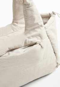 Borsa in tessuto beige chiaro con una superficie liscia e leggermente textured. Presenta un compartimento con zip e due manici robusti.