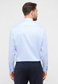 Camicia azzurro chiaro a manica lunga con motivo testurizzato; presenta polsini con bottoni e colletto classico, abbinata a pantaloni scuri.