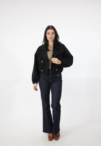 Sort cropped jakke med puffede ærmer og guldknapper, kombineret med mørkeblå flagrende jeans og brune støvler med hæl. Glatte teksturer.