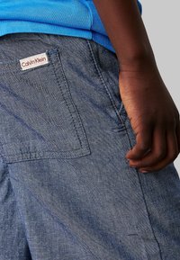 Jasnoniebieskie szorty dżinsowe o fakturowanym wykończeniu, z tylną kieszenią z metką logo Calvin Klein. Ręka spoczywa na pasku.
