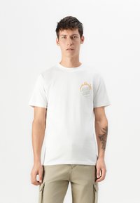 T-shirt de algodão branco com um logótipo gráfico de chamas no lado esquerdo do peito. Design ajustado com mangas curtas e decote redondo clássico.