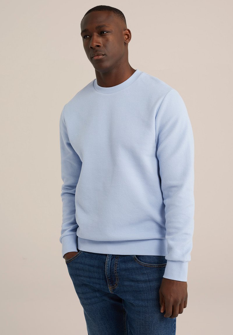 Világoskék, textúrált kapucnis pulóver bordázott mandzsettákkal és aljjal, sötétkék farmernadrággal párosítva, klasszikus környakú fazonnal.