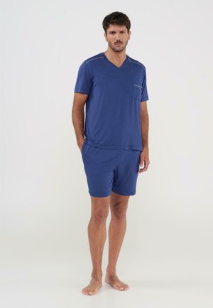 Hombre de pie descalzo, vistiendo una camiseta azul de manga corta con cuello en V y pantalones cortos a juego, frente a un fondo claro y liso.