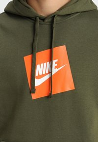 Olivegrön huvtröja med en stor orange Nike-logotyp. Har dragsko för huvan, mjuk textur och en fyrkantig logotypdesign.