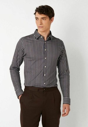 Junger Mann trägt ein schwarz-weiß vertikal gestreiftes Langarmshirt, das in dunkelbraune Hosen gesteckt ist, steht mit den Händen in den Taschen.