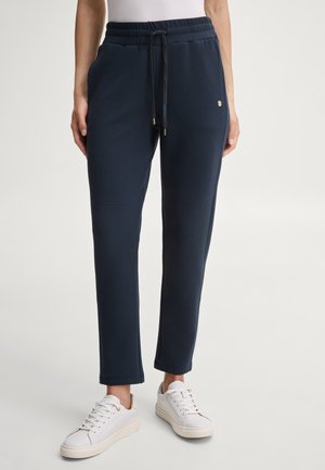 Persona che indossa pantaloni jogger blu navy con coulisse in vita e scarpe da ginnastica bianche, in piedi contro uno sfondo chiaro e semplice.