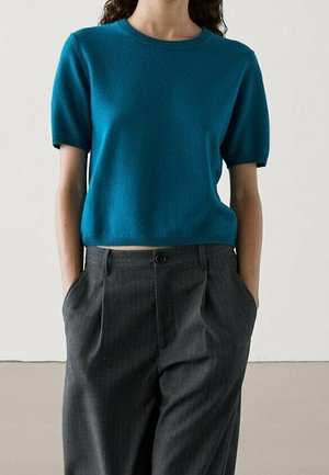 Woman wearing a teal short-sleeve sweater and high-waisted dark gray pinstripe pants with hands in pockets.