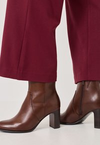 Braune Lederstiefeletten mit einem quadratischen Zehenbereich, Blockabsatz und Reißverschlussdetails, kombiniert mit burgunderfarbenen weit geschnittenen Hosen.