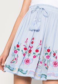 Jasnoniebieska spódnica z tkaniny z haftem kwiatowym w kolorze czerwonym i różowym, z marszczoną talią i szczegółami sznurka z chwostami.