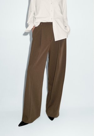 Lichtbruine wijde broek van gladde stof, met een hoge taille en twee voren plooien. Gepaarde met een lichtbeige overhemd met knopen en zwarte hakken.