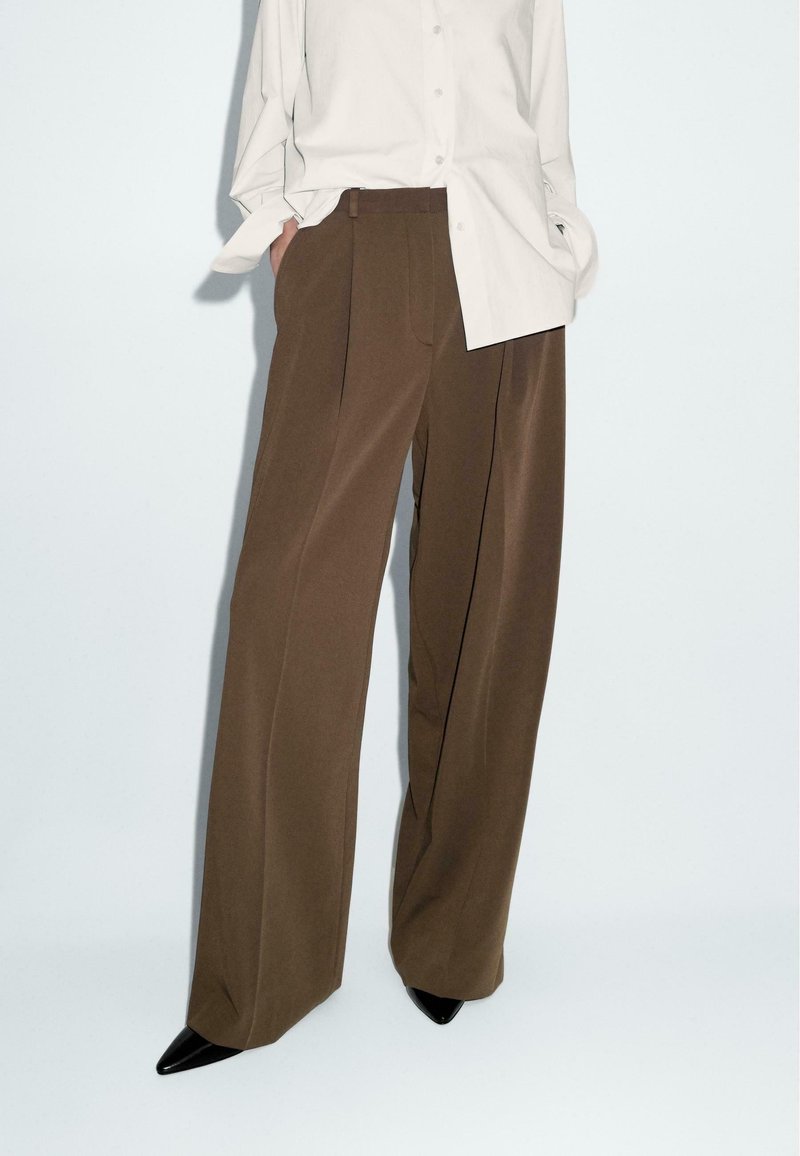 Pantaloni larghi marroni realizzati in tessuto liscio, con vita alta e due pieghe frontali. Abbini a una camicia button-up beige chiaro e tacchi neri.