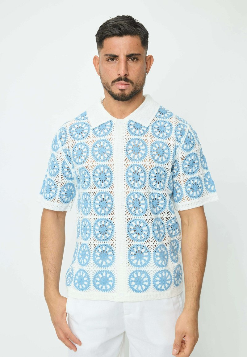 Polo en crochet blanc et bleu clair, affichant des motifs circulaires et des manches courtes, avec un col blanc contrastant et une fermeture à boutons.