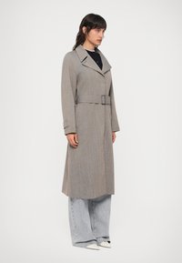 Szary/włókniony płaszcz z zaakcentowanym kołnierzem, z paskiem w talii i bocznymi kieszeniami, w połączeniu z luźnymi, jasnoniebieskimi dżinsami i białymi butami.