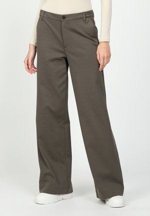 Person iført højtaljede, bredbenede olivengrønne bukser, langærmet beige top og hvide sneakers, står mod en ensfarvet baggrund.