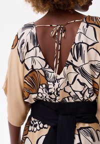 Bloemenprint jurk in crème en bruine tinten, met een diepe V-hals aan de achterkant, losse mouwen en een zwarte tailleband voor een gedefinieerde silhouet.