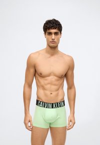 Bez trička, muž s kudrnatými tmavými vlasy, oblečený v světle zelených boxerkách Calvin Klein, stojí proti jednoduchému bílému pozadí.