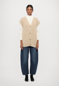 Beige strikket vest med korte ærmer, der har en v-halsudskæring og fem knapper. Bæres over en hvid turtleneck og stylet med brede, mørkeblå jeans.