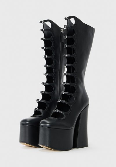 Bottes noires à plateformes, montantes jusqu'aux genoux, avec de hauts talons épais, des bords festonnés et plusieurs petites boucles en argent sur les côtés avant.