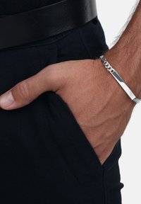 La mano di un uomo in tasca dei pantaloni scuri, indossando un braccialetto d'argento e una cintura nera su uno sfondo chiaro.