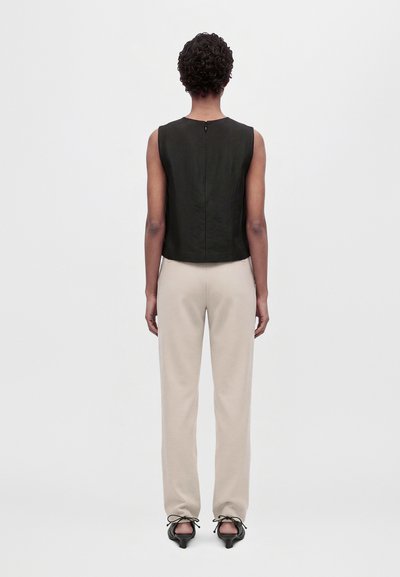 Un mannequin vu de dos porte un haut noir sans manches, un pantalon crème et des chaussures noires à talons sur un fond blanc uni.