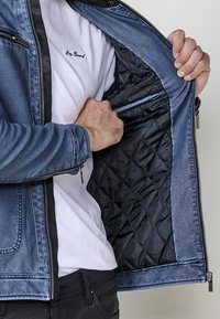 Una persona che indossa una giacca di pelle blu apre la cerniera e sfila la giacca per rivelare un rivestimento interno trapuntato nero e una maglietta bianca sottostante.