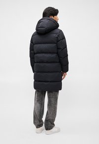 Schwarze gepolsterte Parka mit Kapuze, horizontalen Nähten und einer Länge bis zur Mitte der Oberschenkel. Kombiniert mit grauen Jeans und weißen Turnschuhen.