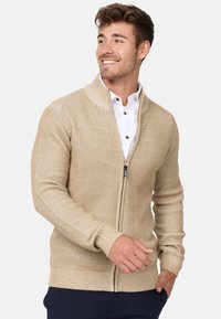 Cardigan beige de cremallera con un patrón de punto texturizado, puños y dobladillo acanalados. Presenta un alto cuello y cremallera frontal para el cierre.
