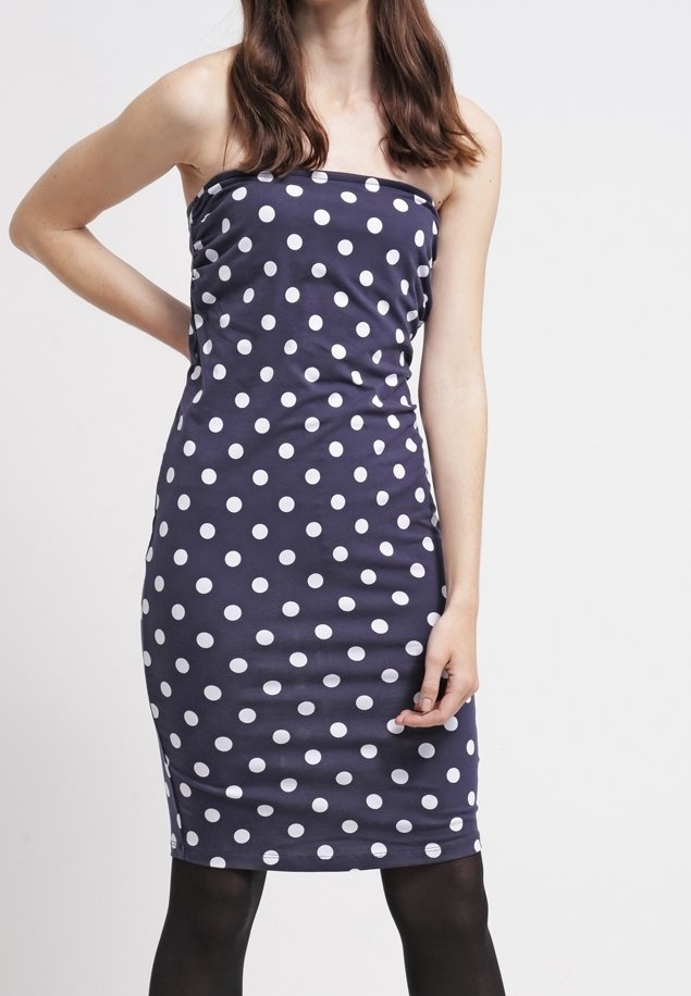 Femme portant une robe bustier bleu marine à pois blancs et des collants noirs, debout devant un fond clair uni.