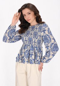 Langärmlige Peplum-Bluse in cremefarbener und blauer Blumenmusterung, mit Rüschen-Details und lockerem Schnitt. Kombiniert mit hellen Hosen.